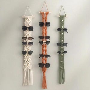 May include: Three vertical wall-hanging organizers made of knotted cord in cream, orange, and olive green. Each organizer features a wooden dowel at the top, intricate macrame patterns, and multiple pairs of sunglasses displayed on the cords.