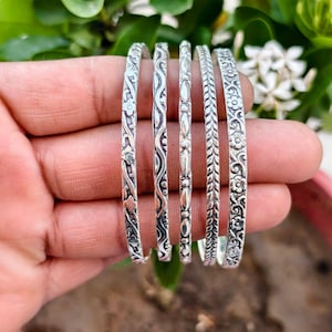 May include: Five silver-toned metal bangles featuring distinct embossed patterns. The designs include floral motifs, wavy lines, a series of small oval shapes, a braided texture, and a detailed vine pattern with small flowers.