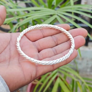 Puede incluir: Un brazalete rígido de color plateado pulido que presenta un diseño continuo de cuerda trenzada. El accesorio circular de metal tiene un acabado metálico reflectante y un grosor uniforme en toda su circunferencia.