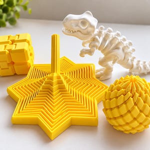 Può includere: Una collezione di oggetti stampati in 3D che include un cubo articolato giallo, una torre pieghevole a forma di stella gialla, una sfera testurizzata gialla e una figura di scheletro di dinosauro articolata bianca.