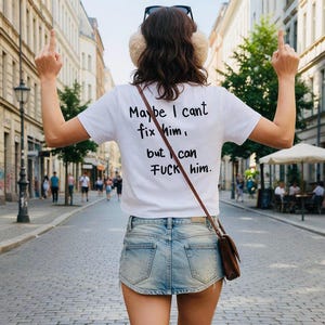 May include: A white short-sleeved t-shirt with black text on the back that reads, "Maybe I cant fix him, but I can FUCK him." The shirt is paired with a light blue denim mini skirt and a brown crossbody bag, worn with beige earmuffs.
