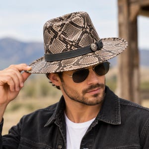 Könnte beinhalten: Ein Fedora-Hut mit einem beigen und schwarzen Schlangenledermuster, akzentuiert durch ein schwarzes Band und ein kreisförmiges silberfarbenes Metall-Concho-Detail. Der Hut hat eine strukturierte Krone und eine breite Krempe mit texturierter Optik.