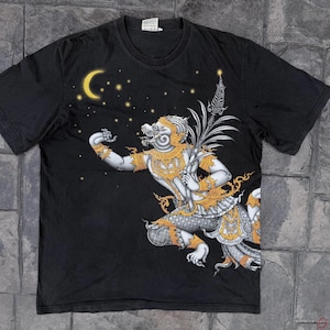 以下が含まれることがあります： 黒の半袖Tシャツ。白と金で描かれた猿のような神話上の人物が木の枝を持っているグラフィックプリントが施されています。デザインには、暗い生地を背景に黄色い三日月と小さな星が含まれています。