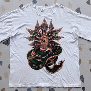 Könnte beinhalten: Ein weißes T-Shirt mit kurzen Ärmeln und einem großen, detaillierten Grafikdruck einer mehrköpfigen grünen Schlange mit verzierten, goldenen, kronenartigen Kopfbedeckungen und einem zentralen kreisförmigen Emblem auf der Brust.