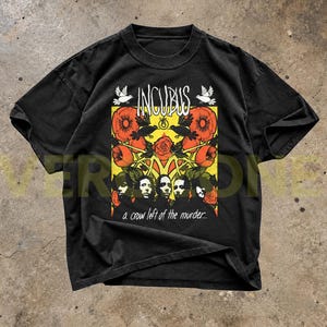 Könnte beinhalten: Ein schwarzes Kurzarm-T-Shirt mit einem Grafikdruck, der den Text INCUBUS und a crow left of the murder zeigt. Das Design umfasst orangefarbene Blumen, schwarze Vögel und fünf Gesichter in einem rechteckigen gelben Rahmen auf der Brust.