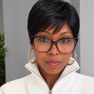 May include: A pair of dark-rimmed, cat-eye glasses with a speckled pattern, worn with large gold hoop earrings and a white, ribbed, high-neck zip-up sweater. The glasses feature a distinct angular frame shape and clear lenses.