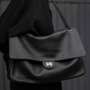 Könnte beinhalten: Eine große schwarze Lederschultertasche mit genarbter Textur, ausgestattet mit einer rechteckigen Klappe, einem silberfarbenen Metalldrehverschluss und einem passenden Schulterriemen. Das Wort Large ist unten rechts in Weiß aufgedruckt.