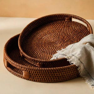 May include: Two round, brown woven rattan trays with raised edges and integrated handles, stacked one inside the other. A light beige, textured fabric cloth with a frayed edge and dark decorative stitching is draped over the side of the trays.