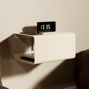 May include: A cream-colored, wall-mounted floating shelf with a curved design. The top surface holds a black digital clock displaying 13:35 and a smartphone. The lower shelf contains a stack of books. The unit is mounted to a plain wall.