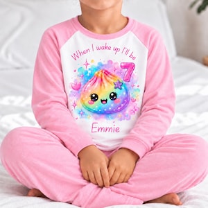 May include: A long-sleeve pyjama set with pink sleeves and trousers, featuring a white centre panel. The graphic shows a rainbow-coloured dumpling character with a smiling face, the number 7, the text "When I wake up I'll be" and the name "Emmie".