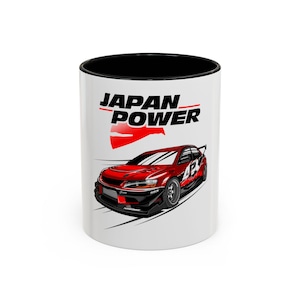 May include: A white ceramic mug with a black interior and rim. The exterior features a graphic of a red sports car with black accents and the text JAPAN POWER in bold black letters above a stylized red sun graphic on a white background.