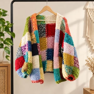 Könnte beinhalten: Eine grob gestrickte Strickjacke mit einem Patchwork-Design aus großen, mehrfarbigen Quadraten in Rot, Gelb, Blau, Grün, Rosa, Weiß und Schwarz. Das Kleidungsstück hat lange, weite Ärmel und eine offene Vorderseite und hängt an einem Holzkleiderbügel.
