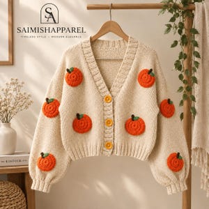 Könnte beinhalten: Eine beigefarbene Strickjacke mit V-Ausschnitt, langen Ärmeln und drei gelben Knöpfen. Das Kleidungsstück ist mit mehreren orangefarbenen, gehäkelten Kürbismotiven verziert, die jeweils einen kleinen grünen Stiel haben und über die Vorderseite und Ärmel verteilt sind.