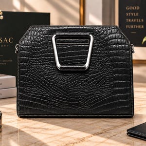 Può includere: Una borsa a mano nera strutturata con una finitura testurizzata effetto coccodrillo e un manico in metallo color argento. La borsa presenta una silhouette trapezoidale con cuciture pulite lungo i bordi e un meccanismo di chiusura sicuro.