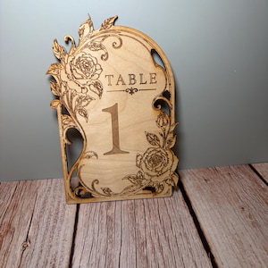 May include: A light-colored wooden table number sign featuring an arched top and intricate laser-cut floral designs. The sign displays the word TABLE above the large numeral 1, with detailed rose engravings on the upper left and lower right corners.