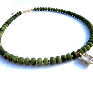 Può includere: Una collana composta da una fila di perline rotonde color verde oliva separate da piccoli distanziatori dorati. Un ciondolo rettangolare in pietra trasparente sfaccettata con montatura dorata pende dalla collana, dotata di una chiusura dorata.