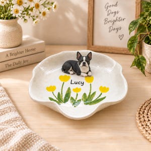 Puede incluir: Un plato de cerámica blanca con borde festoneado, que presenta un diseño pintado a mano de tulipanes amarillos con tallos y hojas verdes. Una pequeña figura tridimensional de un Boston Terrier blanco y negro se sitúa en el centro con el nombre Lucy.