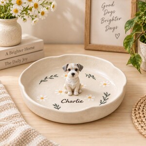 Puede incluir: Un plato decorativo redondo de cerámica color crema con borde festoneado, que presenta una pequeña figura tridimensional de un cachorro beige y blanco en el centro. El plato está decorado con margaritas blancas pintadas, hojas verdes y el nombre Charlie.