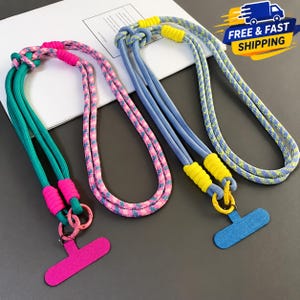 Puede incluir: Dos cordones para teléfono hechos de cuerda trenzada. El cordón izquierdo presenta una cuerda con patrón en verde azulado y rosa con acentos rosa brillante y un conector en forma de T rosa. El cordón derecho presenta una cuerda con patrón en azul y amarillo con acentos amarillos y un conector en forma de T azul.