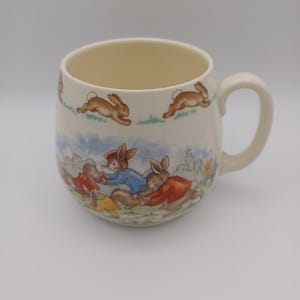 May include: A cream-colored ceramic mug featuring a rounded body and a handle. The exterior is decorated with a repeating pattern of brown rabbits running along the top rim and a colorful illustration of rabbits wearing jackets in a garden scene.