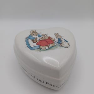 May include: A white heart-shaped ceramic trinket box featuring a colorful illustration of Peter Rabbit characters on the lid. The side of the box displays black printed text that reads, "Cotton-tail and Peter. Once."