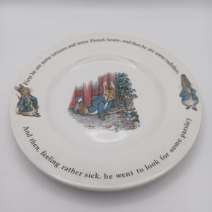 May include: A white ceramic plate featuring illustrations of Peter Rabbit in a blue jacket. The rim displays the text: First he ate some lettuces and some French beans; and then he ate some radishes; And then, feeling rather sick, he went to look for some parsley.