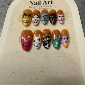 Op de afbeelding: Een set van tien kunstnagels met een amberkleurige basis, elk voorzien van een uniek handgeschilderd kattengezicht in diverse kleuren zoals roze, zwart, wit en blauw. De displaykaart bevat de tekst Nail Art, Thank you for your order.