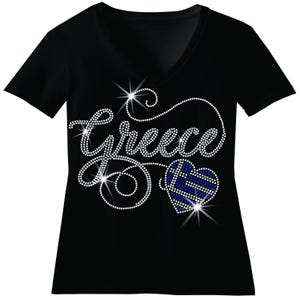 May include: A black V-neck t-shirt featuring the word Greece written in sparkling white rhinestones. The design includes a heart-shaped graphic filled with a blue and white striped pattern, also created with small, reflective rhinestones.