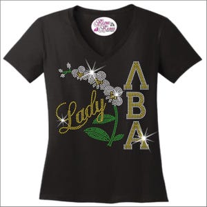 May include: A black V-neck t-shirt featuring a rhinestone design on the front. The graphic includes the word Lady in yellow, a white and yellow orchid flower, green leaves, and the letters ABA in yellow rhinestones. A Bling Babes Boutique logo is visible.