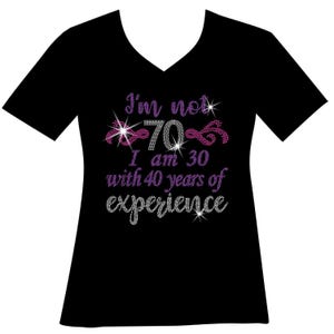 May include: Black V-neck t-shirt with the text "I'm not 70, I am 30 with 40 years of experience" in a rhinestone design. The text is in purple and silver. The number 70 is in silver with pink accents.