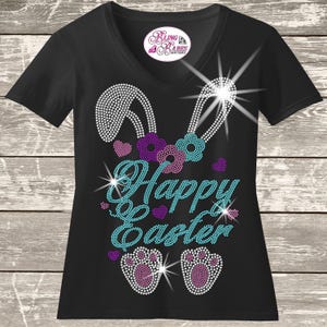 May include: A black V-neck t-shirt featuring a rhinestone design of bunny ears, flowers, and paw prints with the text "Happy Easter" in light blue. The design includes purple and light blue accents, with a Bling Babes Boutique logo at the neck.