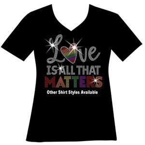 May include: A black V-neck t-shirt featuring a rhinestone graphic that reads Love IS ALL THAT MATTERS, with the word Love incorporating a rainbow-colored heart. The text Other Shirt Styles Available is printed in white at the bottom of the design.