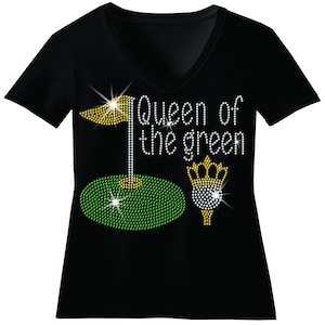 May include: Black V-neck t-shirt with a golf-themed design. The design features a golf flag, a golf ball with a crown, and the text "Queen of the green", all embellished with sparkling rhinestones. The golf elements are in yellow and green.