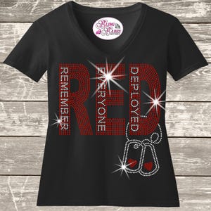 May include: A black V-neck t-shirt featuring a rhinestone graphic that spells RED in large letters, with the words REMEMBER EVERYONE DEPLOYED integrated into the design. Two silver rhinestone dog tags with red hearts hang below the text.