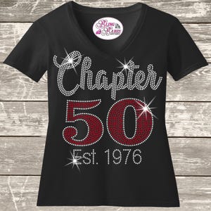 May include: A black V-neck t-shirt featuring a rhinestone design on the front. The text reads Chapter 50 in white and red rhinestones, with Est. 1976 below in white. A Bling Babes Boutique logo is printed on the inside neck label area.