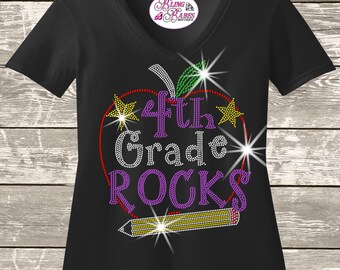 May include: A black V-neck t-shirt featuring a rhinestone graphic of an apple outline, a pencil, two stars, and the text 4th Grade ROCKS. The apple is outlined in red with a green leaf, while the text and pencil are filled with purple, white, and yellow rhinestones.