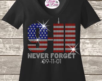 May include: A black V-neck t-shirt featuring a rhinestone graphic of the number 9-11 with the American flag pattern, two vertical towers, and the text NEVER FORGET 09-11-01. The design is embellished with sparkling crystal accents throughout.