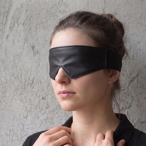 Op de afbeelding: Een zwart lederen blinddoek met een gladde, matte afwerking en een voorgevormde pasvorm die de ogen volledig bedekt. De blinddoek is voorzien van een brede, verstelbare band die rond het hoofd loopt voor een veilige en comfortabele pasvorm.