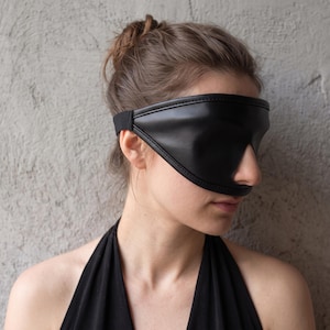Puede incluir: Un antifaz de piel sintética negra con una superficie lisa y contorneada, bordes cosidos y una correa elástica negra flexible. El diseño cubre los ojos por completo manteniendo una forma curva que descansa sobre el puente de la nariz.