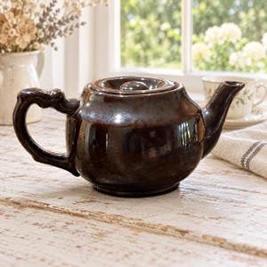 May include: A dark brown glazed ceramic teapot with a rounded body, a short spout, and a curved handle. The teapot features a matching lid with a small knob handle. The surface has a glossy finish that reflects light, showing subtle variations in color.