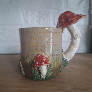 Peut inclure: Une tasse en céramique avec une finition émaillée dans des tons de beige et de vert, dotée d'une anse sculptée en forme de champignon en trois dimensions avec un chapeau rouge à pois blancs. L'extérieur est orné de champignons et de feuillage.