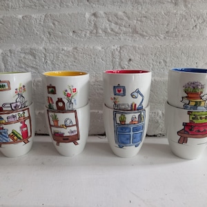 May include: Four white ceramic mugs stacked in pairs, each featuring hand-painted illustrations of furniture and home decor. The interiors are colored in lime green, yellow, pink, and blue. The designs include shelves, drawers, plants, and a cat.