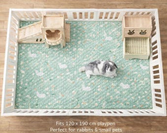 Puede incluir: Un parque rectangular blanco para mascotas con una alfombra verde claro con estampado de conejos blancos. En el interior hay una rampa de madera, una pequeña torre de madera y un comedero beige. El parque se adapta a un área de 120 x 190 cm.
