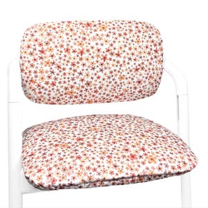 Peut inclure: Une chaise dotée d'une structure blanche et de coussins rembourrés pour l'assise et le dossier. Le tissu présente un motif dense et répétitif de petites fleurs style aquarelle dans des tons orange, rose et marron sur fond blanc.