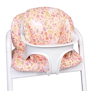 Peut inclure: Une chaise haute blanche dotée d'un coussin de siège rembourré sur fond pêche clair. Le coussin présente un motif répétitif de licornes blanches, de fleurs roses et de feuillages verts. Un support de plateau en plastique blanc est fixé au siège.