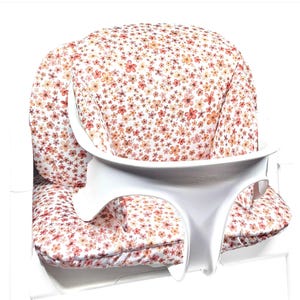 Peut inclure: Un coussin de chaise haute avec une base en tissu blanc orné d'un motif dense et répétitif de petites fleurs orange, roses et rouges. Le coussin est équipé d'un plateau et d'un cadre de support en plastique blanc, avec la marque Cybex visible.