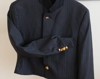 Puede incluir: Una chaqueta azul marino oscuro con rayas diplomáticas, cuello mao y botones dorados. La prenda presenta una silueta estructurada con mangas largas, cada una terminada con una hilera de botones dorados decorativos en los puños.