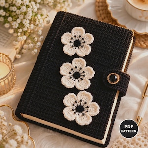 May include: A black crocheted notebook cover featuring three cream-colored crocheted flowers with black centers arranged vertically. The cover includes a side strap with a circular black and gold button closure. A black circular icon with the text PDF PATTERN is visible.