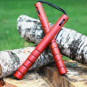May include: A pair of red wooden nunchucks featuring a smooth, polished finish with distinct horizontal grooves along the length of each handle. The two handles are connected at the top by a thin, black cord threaded through oval-shaped openings.