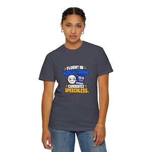 May include: A charcoal grey short-sleeve t-shirt featuring a graphic with the text "FLUENT IN MEDICAL CODING. CURRENTLY SPEECHLESS." The design includes a white cartoon face with a neutral expression and a blue book labelled "ICD-10" in white.